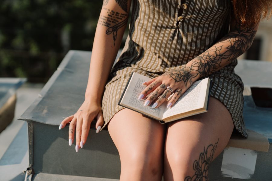 A light-skinned woman with tattoos on her arms, hands, and leg sits with a book open in her lap.