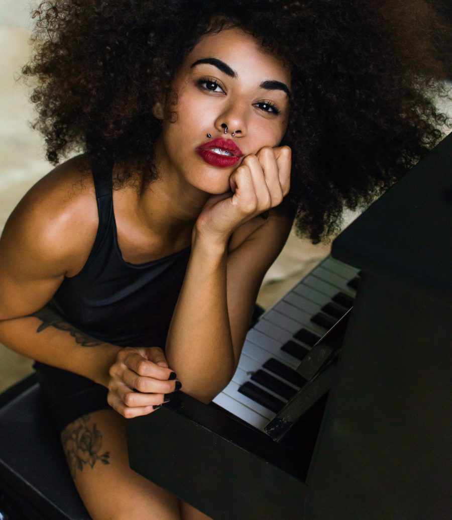 A dark-skinned woman with red lipstick sits at the edge of a piano, with her chin resting in her hand and her elbow on the keys.
